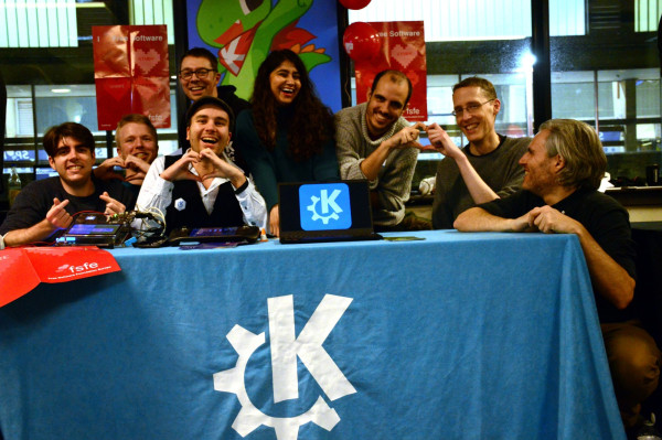 KDE members smile and laugh while making the heart sign with their hands at the KDE booth at FOSDEM 2023.