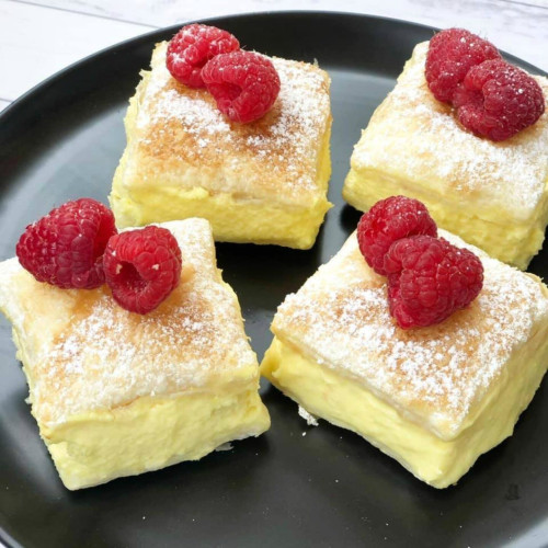 Four squares of vanilla slice. A thick, whipped vanilla custard sandwiched between puff pastry, dusted with powdered sugar, and topped with a pair of raspberries.
