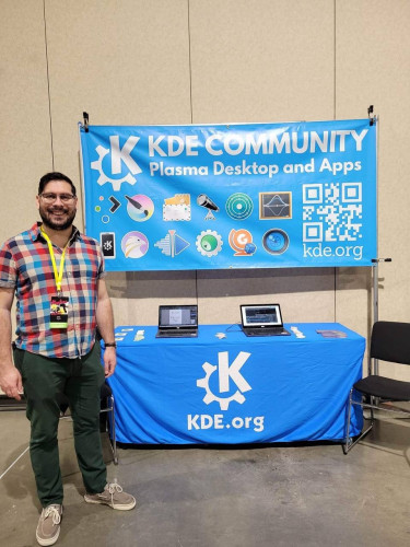Drew is standing in front of a KDE booth where you can see a banner displaying the icons of several KDE apps, and a table with two laptops and stickers.
