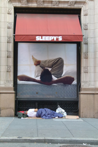 Unhoused person sleeping on a cardboard mat under a window sign ad of someone sleeping on a comfortable mattress.