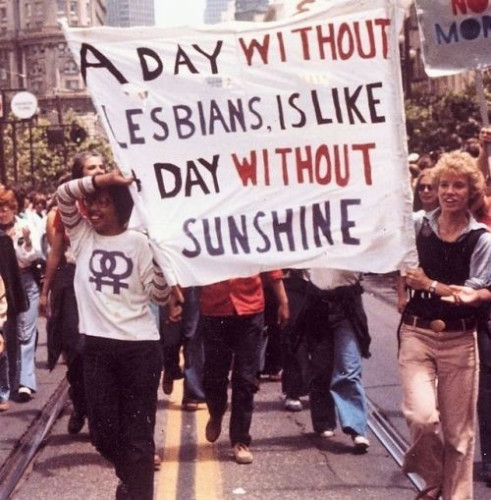 A cloth sign with hand painted lettering saying 'A DAY WITHOUT LESBIANS IS LIKE A DAY WITHOUT SUNSHINE' — a callback to a marketing slogan advocated by Anita Bryant to promote orange juice.