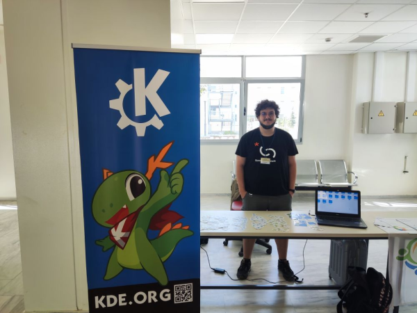 George standing at the KDE stand at FOSSCOMM. There are stickers and a laptop on the table.