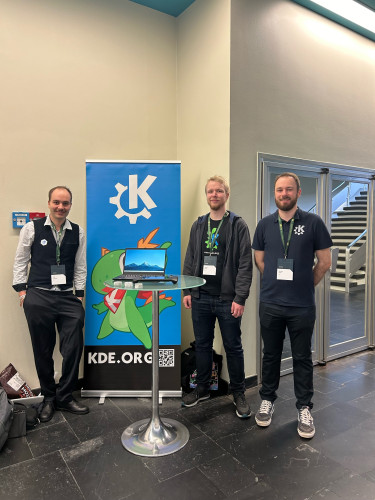 From left to right, Carl Schwan and Tobias and Nicolas Fella, standing around a tall table with a KDE-powered laptop and the Steam Deck. Behind them you can see a standup banner with the KDE logo, Konqi the dragon and a link to KDE's website https://kde.org.
