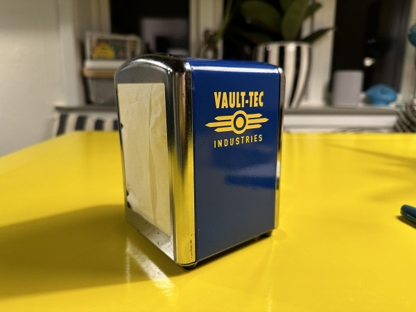 Napkin dispenser, blue with yellow text and logo spelling Vault-Tec Industries.